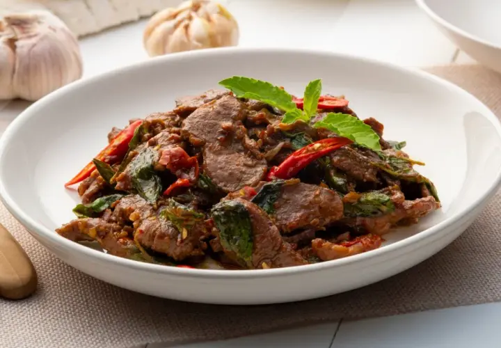 Aksamitne kawałki wołowiny w aromatycznym sosie, z dodatkiem świeżej bazylii i papryczek chili. Pyszne wołowina po tajsku na białym talerzu.
