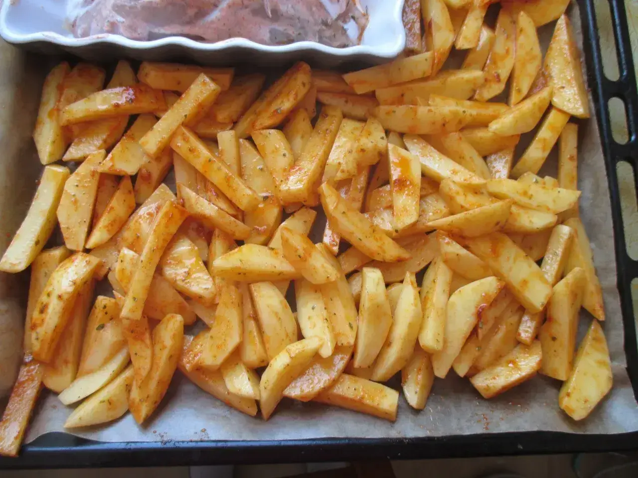 Gotowe do pieczenia frytki w piekarniku, pokrojone w łódeczki i doprawione przyprawami, leżą na blasze wyłożonej papierem.