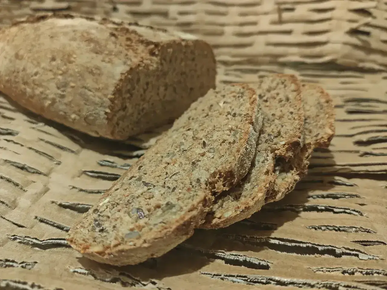 Chleb orkiszowy, pokrojony w kromki, z widocznymi ziarnami, leży na falistej tekturze.